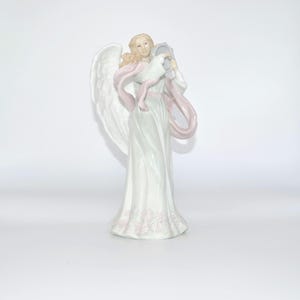 May include: A white porcelain angel figurine with pink wings and a pink ribbon holding a small white porcelain baby. The angel is wearing a white dress with pink floral details.