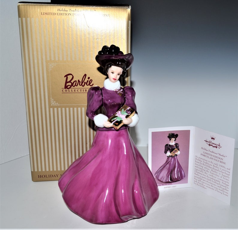 May include: A porcelain figurine of a Barbie doll dressed in a purple gown and a black hat, holding a book. The figurine is part of the Hallmark Traditions Barbie Collectibles series.