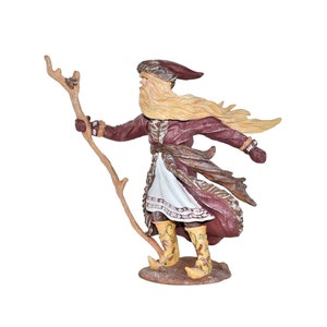 May include: A figurine of a bearded man wearing a brown and white fur-trimmed robe, a red hat, and yellow boots. He is holding a stick in his right hand and his left hand is raised. The figurine is standing on a brown base.