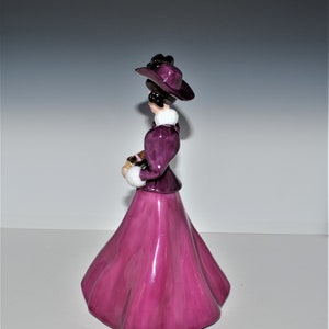 May include: A porcelain figurine of a woman wearing a purple dress and hat. She is holding a small book in her hands.