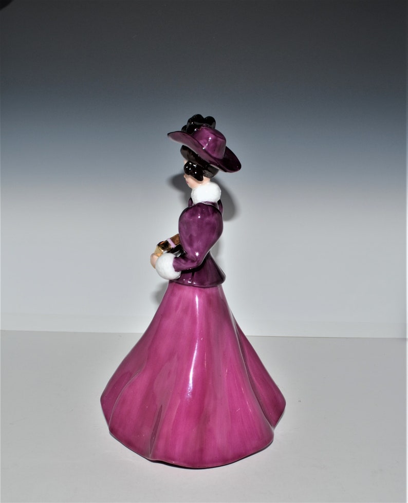 May include: A porcelain figurine of a woman wearing a purple dress and hat. She is holding a small book in her hands.