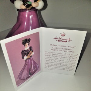 May include: A porcelain figurine of a Barbie doll dressed in a purple dress and a black hat, holding a book. The figurine is part of the Hallmark Holiday Traditions collection and is dated 1907.