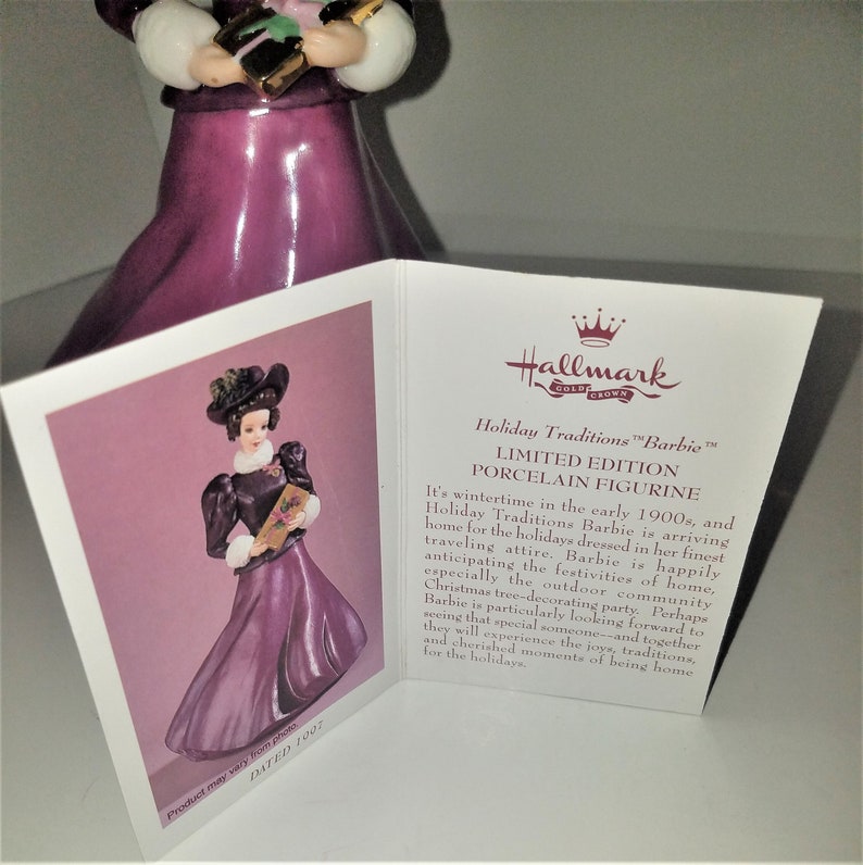 May include: A porcelain figurine of a Barbie doll dressed in a purple dress and a black hat, holding a book. The figurine is part of the Hallmark Holiday Traditions collection and is dated 1907.