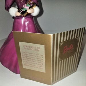 May include: A certificate of authenticity card for a Barbie Holiday Homecoming Collection porcelain figurine. The card is gold with a striped pattern and features the Barbie logo. The text on the card reads "Certificate of Authenticity" and "This card certifies that this porcelain figurine is one of a limited production of no more than 24,500 pieces. Based on the Hallmark Special Edition Holiday Traditions Barbie doll, this figurine is a part of the Holiday Homecoming Collection."