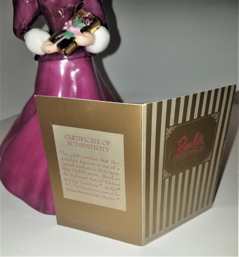 May include: A certificate of authenticity card for a Barbie Holiday Homecoming Collection porcelain figurine. The card is gold with a striped pattern and features the Barbie logo. The text on the card reads "Certificate of Authenticity" and "This card certifies that this porcelain figurine is one of a limited production of no more than 24,500 pieces. Based on the Hallmark Special Edition Holiday Traditions Barbie doll, this figurine is a part of the Holiday Homecoming Collection."