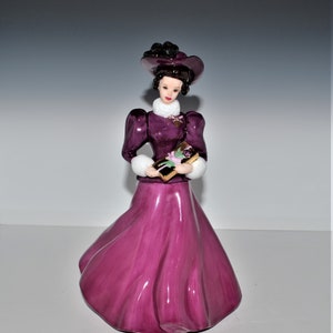 May include: A porcelain figurine of a woman in a purple dress and hat, holding a wrapped gift box with a floral design. The figurine has a white fur trim on the sleeves and a delicate gold trim on the dress.