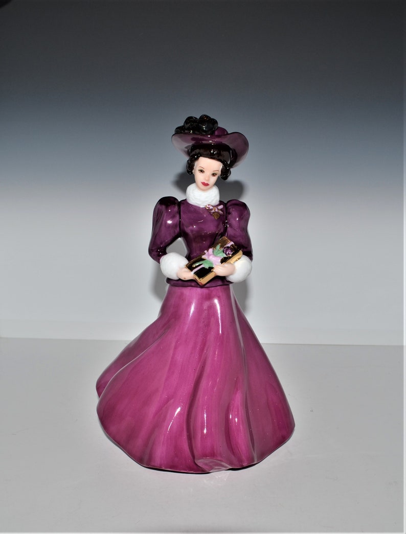 May include: A porcelain figurine of a woman in a purple dress and hat, holding a wrapped gift box with a floral design. The figurine has a white fur trim on the sleeves and a delicate gold trim on the dress.