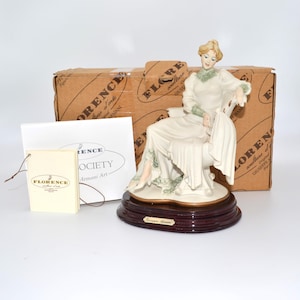 May include: A porcelain figurine of a seated woman in a white dress with green accents, resting on a chair. The figurine is on a dark wooden base. The box behind the figurine reads "Florence" and "Armani Art".