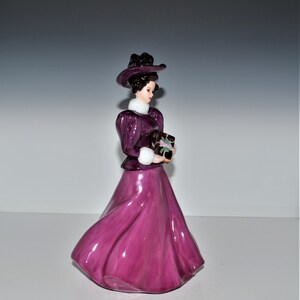 May include: A porcelain figurine of a woman in a purple dress and hat, holding a package. She is wearing a white fur collar and has dark hair.
