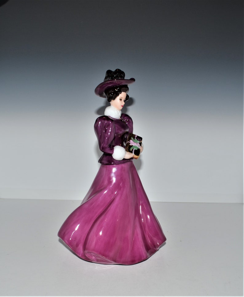 May include: A porcelain figurine of a woman in a purple dress and hat, holding a package. She is wearing a white fur collar and has dark hair.