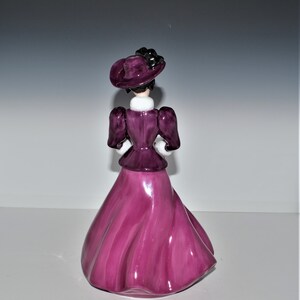May include: A ceramic figurine of a woman wearing a purple dress and hat. The dress has a flowing skirt and the hat has a black ribbon.