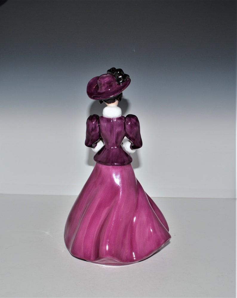 May include: A ceramic figurine of a woman wearing a purple dress and hat. The dress has a flowing skirt and the hat has a black ribbon.