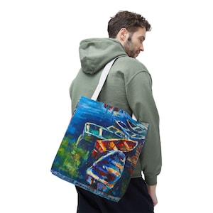May include: A blue tote bag with a colorful abstract painting of boats. The painting features shades of blue, red, orange, and green. The bag has a white strap and is carried over the shoulder.