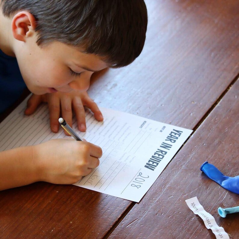 May include: A young person sits at a wooden table, writing on a white sheet of paper with a black pen. The paper has the title "Year in Review 2018" printed on it. There are two blue balloons and a piece of paper with the text "100m fastest & sons" on the table.