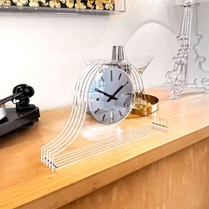 Könnte beinhalten: Eine moderne, transparente Acryluhr mit silbernem Zifferblatt und schwarzen Zeigern. Die Uhr hat ein einzigartiges Design mit gebogenen Acrylhaltern. Sie steht auf einer hellen Holzoberfläche, mit anderen dekorativen Gegenständen im Hintergrund.