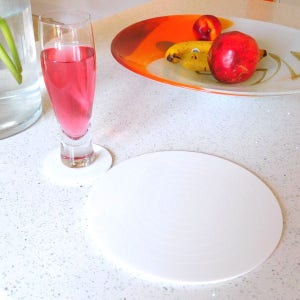 May include: Two white round coasters with a concentric circle design. One coaster is under a glass of pink liquid. The other coaster is on a white countertop.