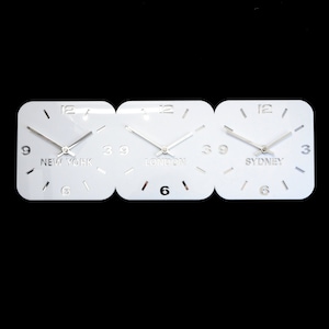 May include: A white wall clock with three square faces, each displaying a different time zone: New York, London, and Sydney. The clock hands are silver, and the numbers are cut out of the white surface.