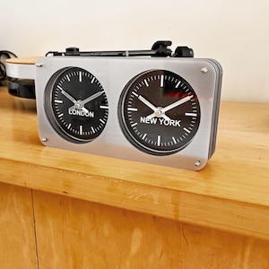 May include: A silver-colored dual-dial clock displaying the time for London and New York. The clock has a rectangular frame with two circular black clock faces. The word "LONDON" is printed on the left clock face, and "NEW YORK" on the right.
