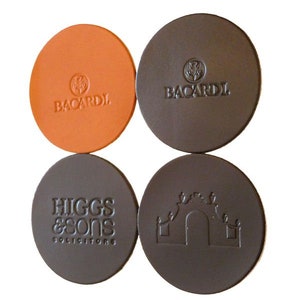 May include: Four round leather coasters with different designs. One coaster is orange with the Bacardi logo, another is brown with the Bacardi logo, a third is brown with the text "Higgs & Sons Solicitors", and the last is brown with a stylized building design.