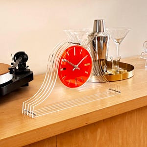 May include: A retro-style red clock with a clear acrylic frame and white lines. The clock is on a wooden surface, next to a cocktail shaker, martini glasses, and a record player. The clock's face has white hands and markers.