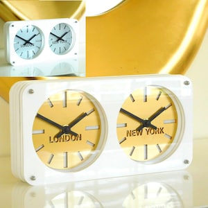 May include: White dual-time clock with gold-colored clock faces. The clock faces display "LONDON" and "NEW YORK" with black hands.