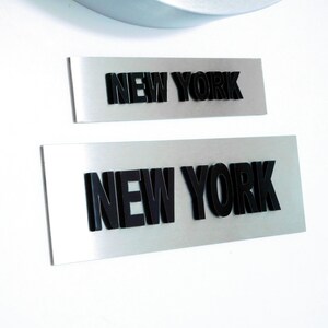 Könnte beinhalten: Zwei silberne Metallschilder mit dem Text "NEW YORK" in schwarzen erhabenen Buchstaben. Das obere Schild ist kleiner als das untere Schild.