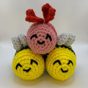 May include: Three crocheted stuffed toys: a pink bee with red wings, and two yellow bees with gray wings. All three have smiling faces.