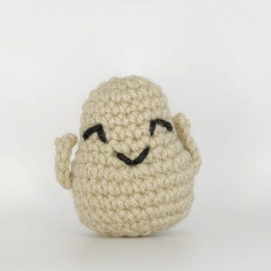 May include: A crocheted, off-white plush toy shaped like a smiling character. The toy has a rounded body, small arms, and a stitched black face with a curved mouth and arched eyebrows. The background is plain white.