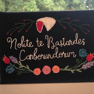 May include: A black canvas with white embroidery that reads "Nolite te Bastardes Carborundorum" surrounded by a floral wreath with red, pink, and blue flowers.