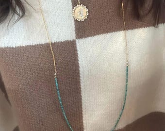 May include: A gold necklace featuring a turquoise bead strand and a pendant with a floral design. The necklace is layered over a second gold chain with a circular pendant. The background is a brown and white checkered sweater.