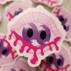 以下が含まれることがあります： ピンクのまぶたにピンクの液体が滴る、漫画の目のステッカーセット。目は白く光る紫です。