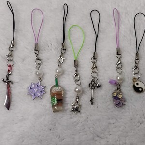 Peut inclure: Collection de sept breloques décoratives pour téléphone. Chaque breloque présente un pendentif différent : une épée, un flocon de neige, une tasse de café, une tortue, une clé, une abeille et un symbole yin-yang. Les breloques ont des attaches en métal argenté et des accents colorés.
