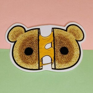 Op de afbeelding: Een sticker van een cartoon gegrilde kaassandwich met een stroperige kaas die eruit druipt. De sandwich is gevormd als een beer met twee ronde ogen.