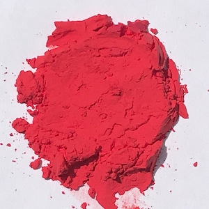 May include: A pile of bright red powder on a white surface. The powder is finely ground and has a smooth texture.