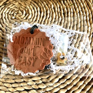 May include: A terracotta-colored ceramic air freshener with the text "Mommin' ain't easy" in white lettering. The air freshener is packaged with a small glass bottle of essential oil.