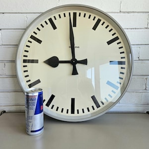 Può includere: Un grande orologio rotondo con un quadrante color crema e lancette e indicatori neri. L'orologio ha la scritta "SIEMENS" sul quadrante. Una lattina d'argento di Red Bull è accanto all'orologio.