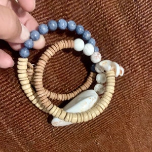 Pu&ograve; includere: Tre braccialetti sovrapposti con perline blu, bianche e marroni. I braccialetti sono fatti di legno e conchiglia.