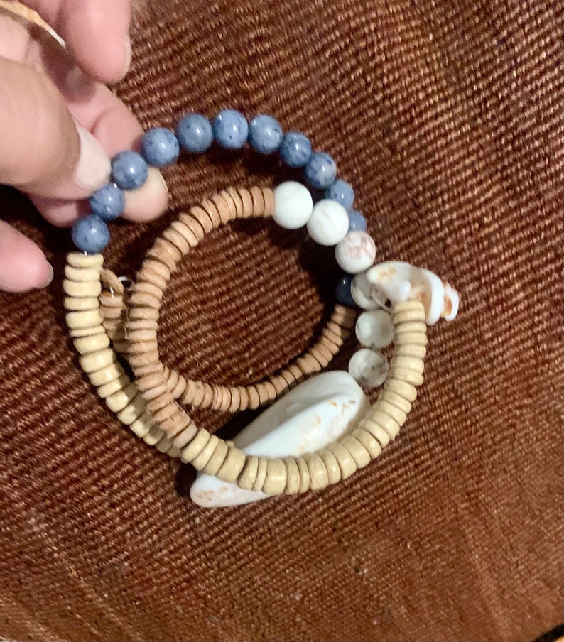 Pu&ograve; includere: Tre braccialetti sovrapposti con perline blu, bianche e marroni. I braccialetti sono fatti di legno e conchiglia.