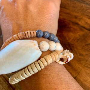 Pu&ograve; includere: Un set di braccialetti con materiali naturali, tra cui perline di legno, pietre bianche e blu e una grande conchiglia bianca.
