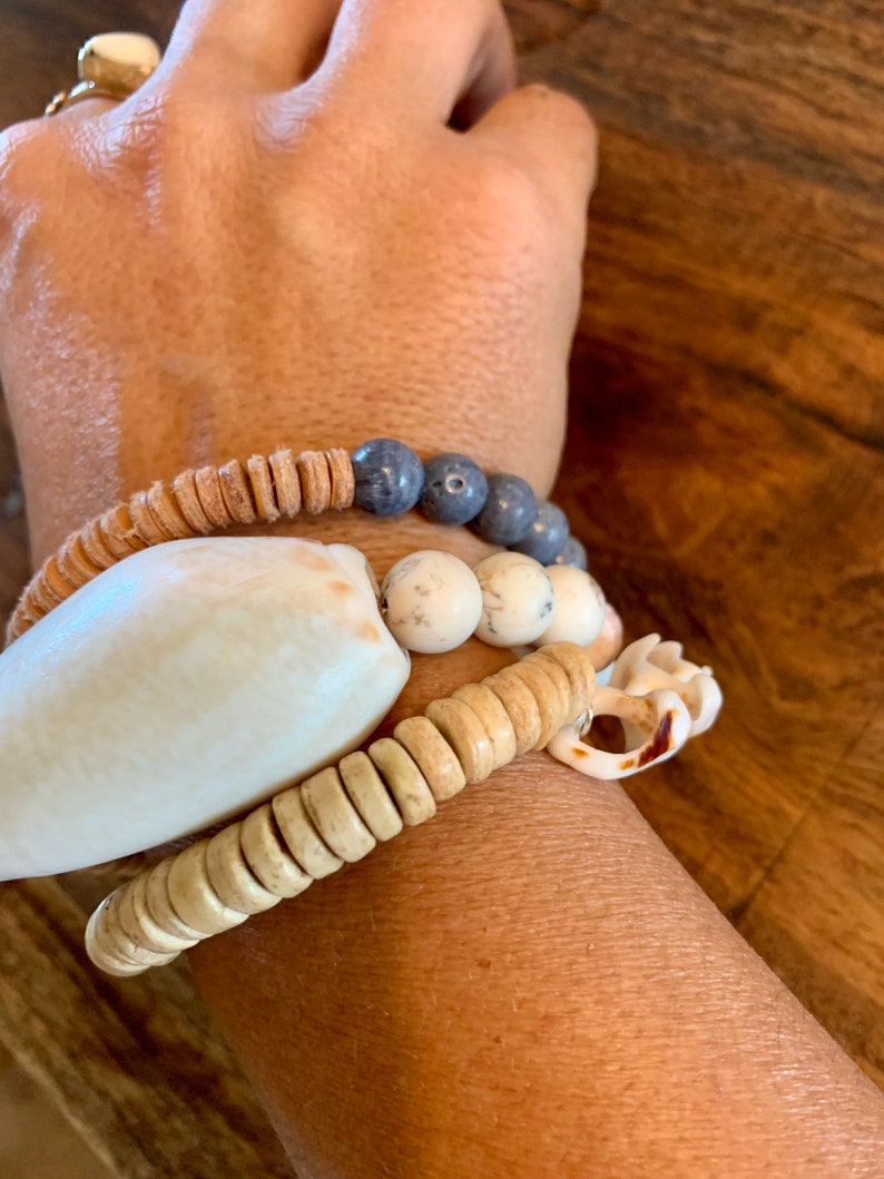 Pu&ograve; includere: Un set di braccialetti con materiali naturali, tra cui perline di legno, pietre bianche e blu e una grande conchiglia bianca.