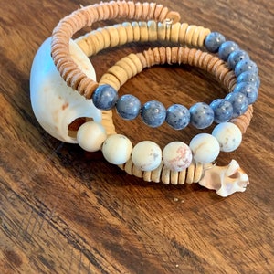 Pu&ograve; includere: Tre braccialetti sovrapposti con perline blu, bianche e marroni. I braccialetti sono realizzati con materiali naturali e hanno uno stile rustico e boh&eacute;mien.