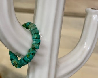 Può includere: Un anello di perline blu turchese fatto di piccole pietre di forma irregolare. L'anello è esposto su un cactus in ceramica bianca.