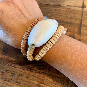 Pu&ograve; includere: Un braccialetto realizzato con materiali naturali, con un grande perno di conchiglia bianca e due strati di perline di legno marrone.