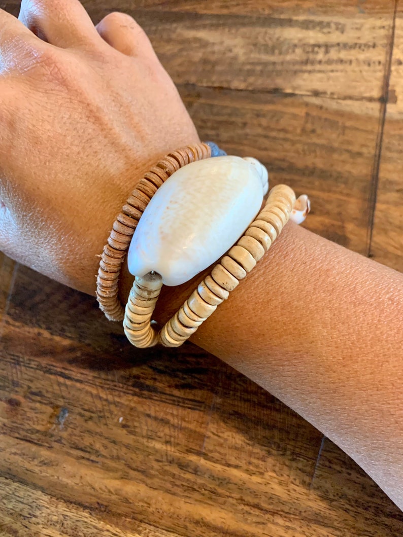 Pu&ograve; includere: Un braccialetto realizzato con materiali naturali, con un grande perno di conchiglia bianca e due strati di perline di legno marrone.