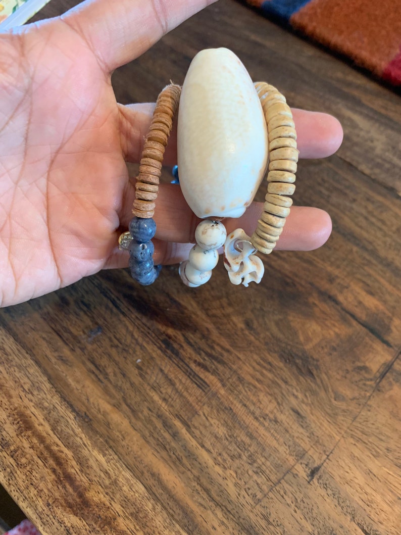 Pu&ograve; includere: Tre braccialetti di perline con un ciondolo a forma di conchiglia bianca. I braccialetti sono fatti di perline marroni, beige e blu.