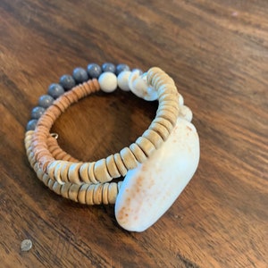 Pu&ograve; includere: Un braccialetto a strati realizzato con perline di legno, perline grigie e una conchiglia bianca. Il braccialetto &egrave; su una superficie di legno.