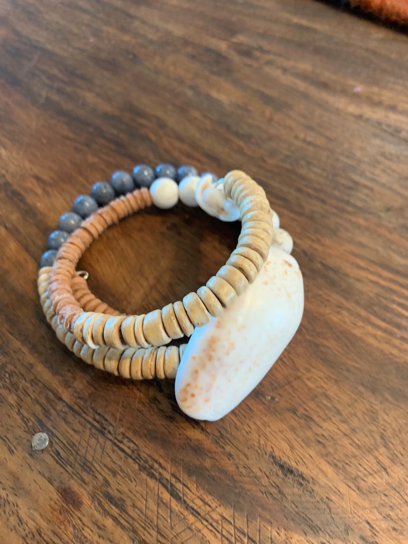 Pu&ograve; includere: Un braccialetto a strati realizzato con perline di legno, perline grigie e una conchiglia bianca. Il braccialetto &egrave; su una superficie di legno.