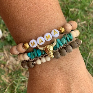 May include: A stack of bracelets featuring a gold bull head charm. The top bracelet has white beads with the word "MAMA" spelled out in gold letters. The other bracelets are made of wood and turquoise beads.