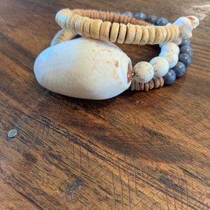 Pu&ograve; includere: Un set di due braccialetti realizzati con materiali naturali. Il braccialetto superiore &egrave; fatto di perline di legno marrone chiaro. Il braccialetto inferiore &egrave; fatto di perline bianche e grigie con un grande ciondolo a conchiglia bianca.
