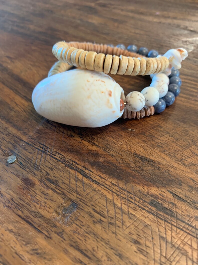 Pu&ograve; includere: Un set di due braccialetti realizzati con materiali naturali. Il braccialetto superiore &egrave; fatto di perline di legno marrone chiaro. Il braccialetto inferiore &egrave; fatto di perline bianche e grigie con un grande ciondolo a conchiglia bianca.
